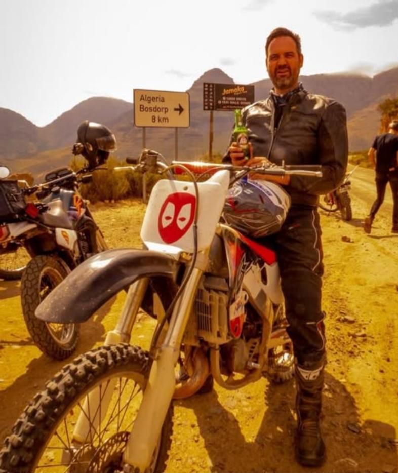 Edrich in riding leathers on a white enduro bike beside a road sign reading Algeria, Bosdorp 8 km, with mountains behind