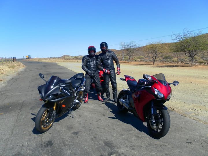 Two riders in black leathers and helmets standing between a black superbike and a red superbike on a dry country road under a clear blue sky